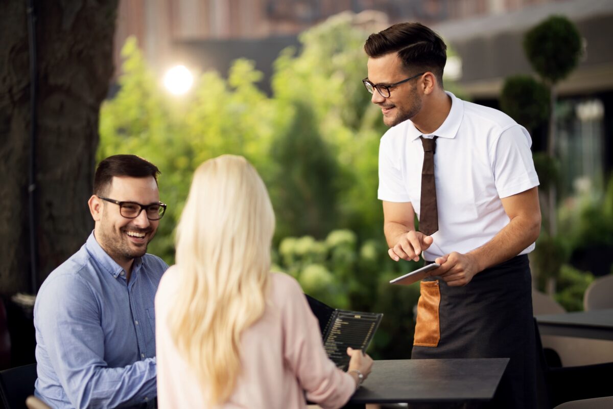 Smiling waiter using touchpad while talking with guests and taking their order